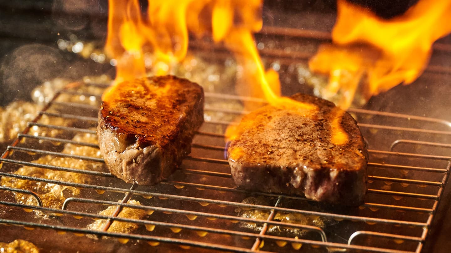Alt設定値：直火の炎に包まれながら網焼きされるステーキ肉と、激しく弾ける脂のシズル感あふれるライブ撮影カット