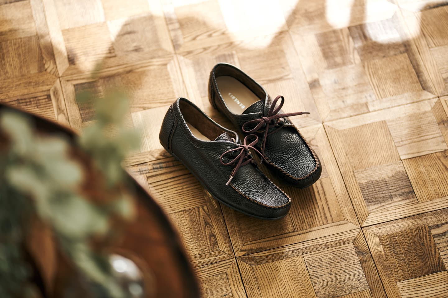大阪のスタジオで撮影された、アンティーク調の寄木細工の床に置かれた黒のレザースニーカー。自然光が差し込む上質な室内空間を演出した、D2Cブランド向けの商品スタイリング写真。