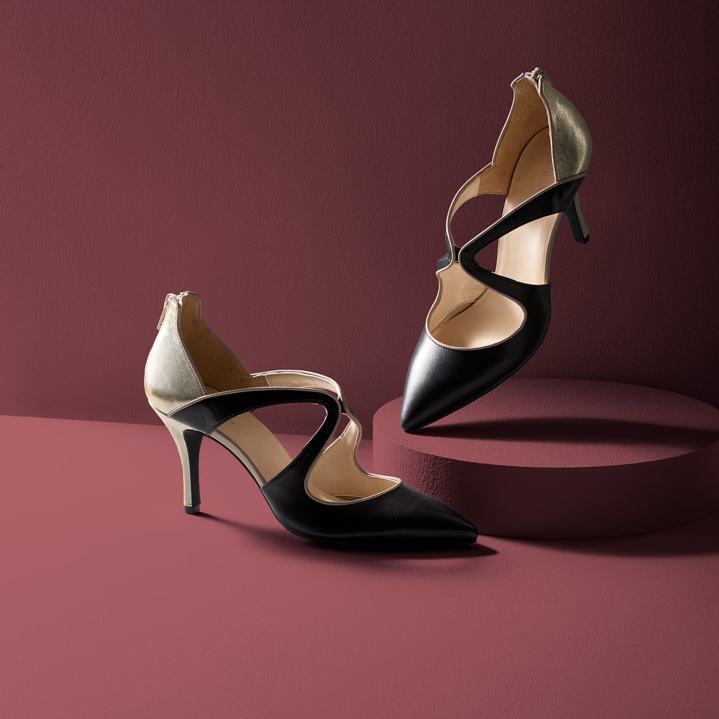 大阪のスタジオで撮影された、深いエンジ色の背景に佇む黒とゴールドのパンプス。円柱の什器を用いた立体的なレイアウトと、サイドからの光で曲線美と素材のコントラストを際立たせた高品位な広告写真。